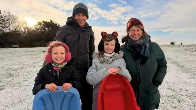 Two grandparents stand with their two grandchildren who are holding sledges, against a snowy background