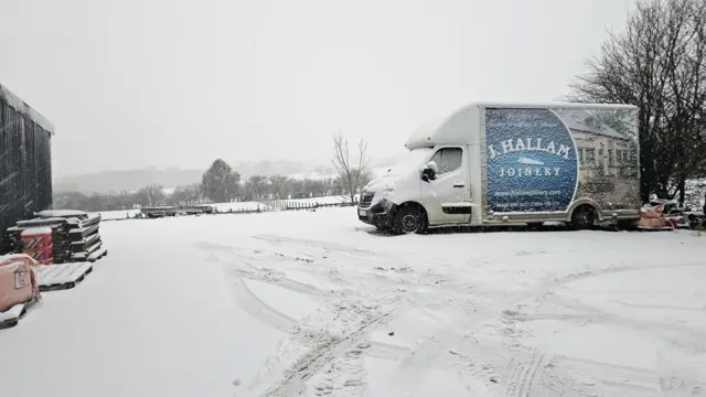 A white van branded with “ J HALLAM JOINERY” is parked on a snow-covered surface. The van features a logo and an image of a house on its side. Snow blankets the ground, with visible tyre tracks and a stack of wooden pallets to the left. Construction materials, partially covered in snow, are scattered nearby.