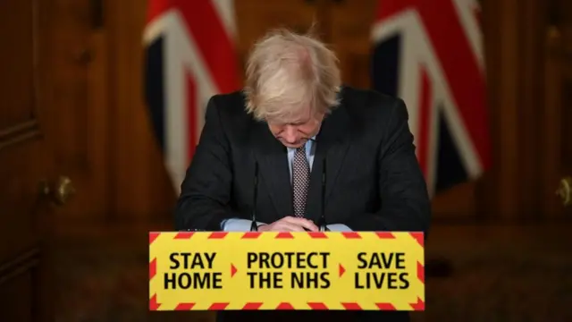 Prime Minister Boris Johnson looks down at the podium during a media briefing, with the words: Stay home, protect the NHS, save lives on the podium
