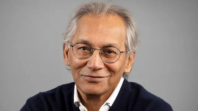 BBC Chair Samir Shah, looking into the camera wearing glasses, white shirt and vneck jumper against grey backdrop