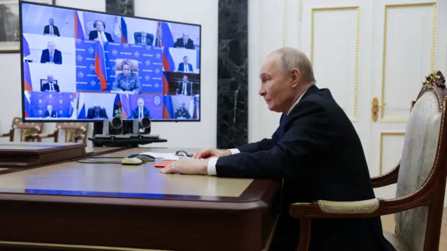 Vladimir Putin in a black suit sits at a wooden desk, a screen split into 13 mini-windows showing members of his security council stands on a separate desk to his right