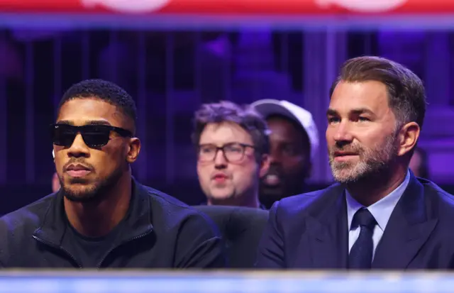 Anthony Joshua is wearing sunglasses and sitting next to Eddie Hearn