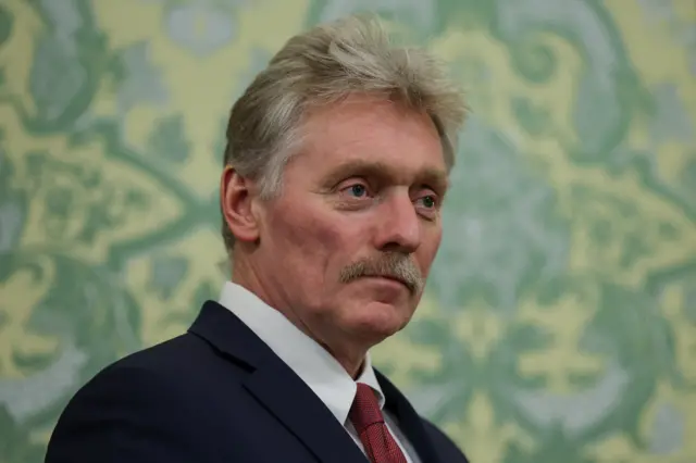 Dmitry Peskov close up. He's wearing a dark suit, white shirt and red tie. Behind him is a wall covered in yellow, green and blue floral brocade