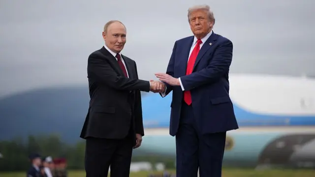 Putin (left) shakes hands with Trump (right). Trump wears a blue suit and red tie, Putin wears a black suit. US Air Force One can be seen behind them