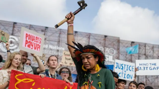 A woman wearing indigenous head-dress raises an axe in the foreground of a crowd protesting in front of the COP30 summit in Belém, Brazil.