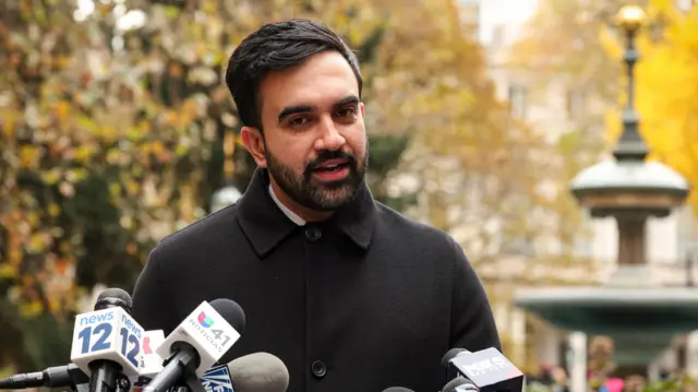 Zohran Mamdani speaks into several mics during an outdoor press conference at City Hall Park. He's wearing a buttoned-up dark coat