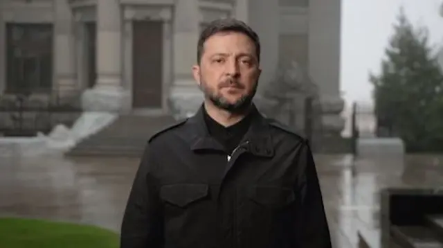 Volodymyr Zelensky stands in a black jacket looking at the camera. He has short dark hair and a beard