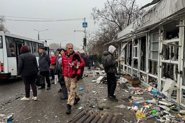 A man in a red jacket carrying a dog observes destroyed shopping pavillions as he walks past