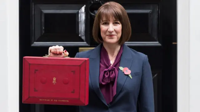 Chancellor Rachel Reeves, a white lady with brown hair in a bob, stands outside Downing Street in a purple shirt and dark blue blazer holding a red briefcase.
