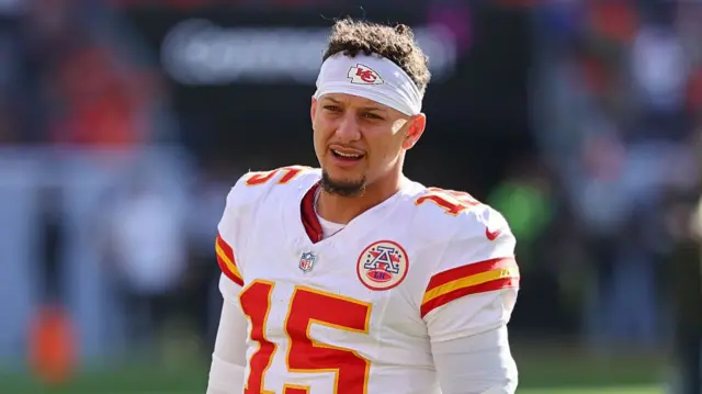 Patrick Mahomes stands on the field wearing a Chiefs away jersey and white Chiefs headband