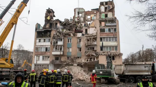 A high rise building completey destroyed, with emergency service workers looking on at the rubble