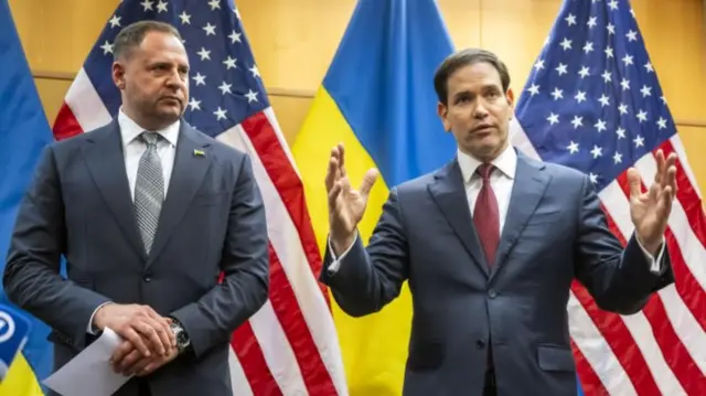 Yermak stands next to and looks at Rubio, who is gesturing with his hands as he speaks. The two men are standing in front of US and Ukrainian flags
