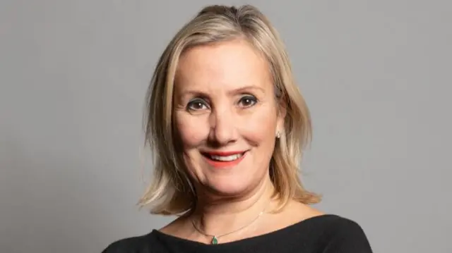 Committee chair Caroline Dinenage has blonde, shoulder-length hair and a black top. She is photographed smiling against a grey background.
