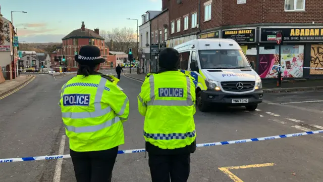 Two police officers in high-visibility jackets stand near a cordoned-off street, facing a white police van with flashing lights.
