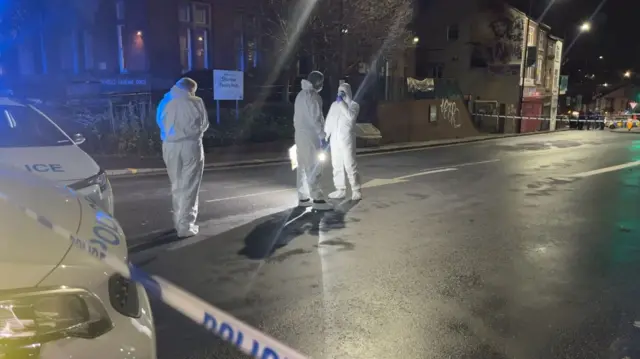 Crime scene investigators in white hazmat suits stand inside a cordoned area of a residential street.