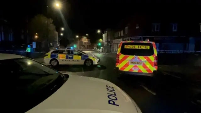 Police cars and a van park next to a cordon at night.