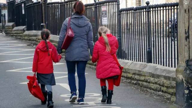 Two young girls wearing red coats hold hands with a woman wearing a grey coat