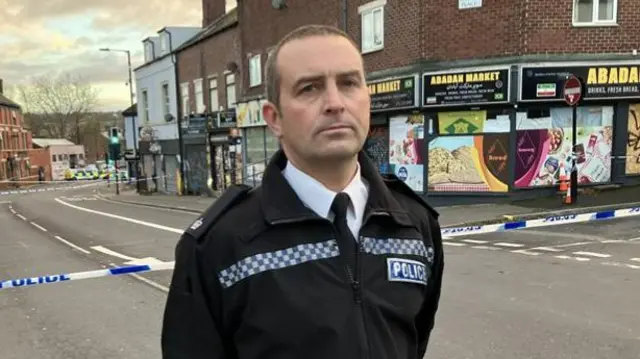 A police officer in a black uniform stands in front of a cordoned-off urban street.