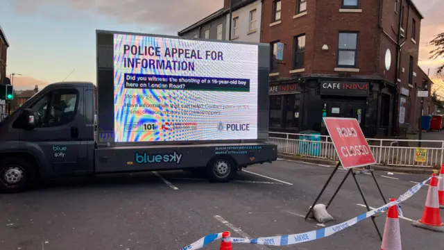 Police appeal billboard on a truck seeks witnesses to a teen’s shooting on London Road. Road is closed with cones, tape, and signage; café and urban buildings in background.