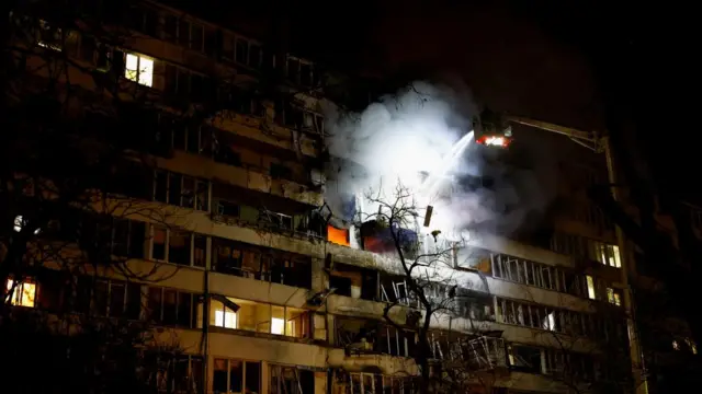 A damaged residential building with windows falling off and smoke emitting from the middle