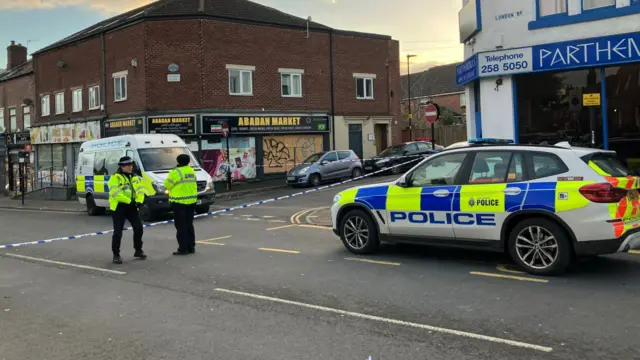 A cordoned-off street scene shows two police officers in high-visibility jackets standing near blue-and-white police tape. A marked police van and SUV with flashing lights are parked nearby. The area appears quiet and secured, with shops including “Abadan Market” visible.