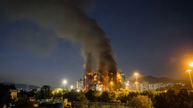 Wide shot showing several buildings of the Wang Fuk Court complex on fire, black and grey smoke billowing in the early night sky