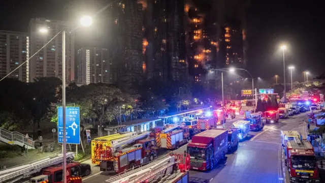 Fire trucks and other emergency vehicles line the highway next to the fire, which glows in the night.