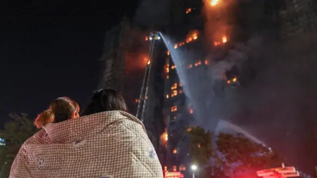 Two people huddle under a blanket, watching fire fighters tackle the Tai Po blaze