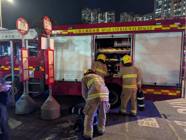 Firefighters take out oxygen masks from fire engines as they prepare to tackle fire. They're wearing yellow uniforms and helmets