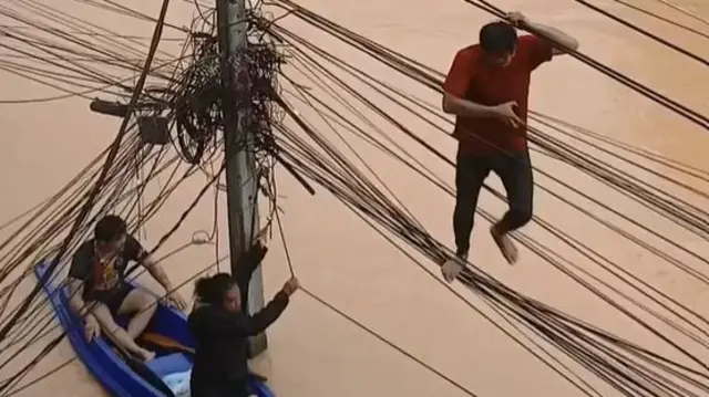 A screengrab showing a man using overhead cables like a rope bridge to get along a flooded street while two men in a blue plastic dinghy use the same cables to pull themselves through the water