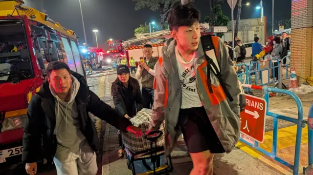 A group of young people pull a trolley filled to the brim with bags as they walk past several fire engines