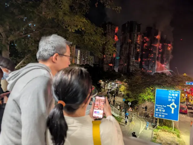 An elderly man (L) stands next to a woman with black hair in a ponytail (R) looking at her phone. They're standing on a high rise part of the street watching three buildings burn in the background