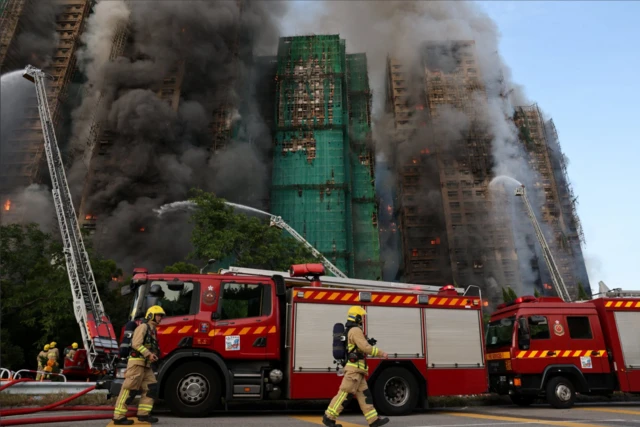 Firefighters in yellow uniforms and helmets walk past a red fire engine parked at the bottom of a green net and bamboo scaffolding-covered building (C). The central building and two other tower blocks to its side are on fire