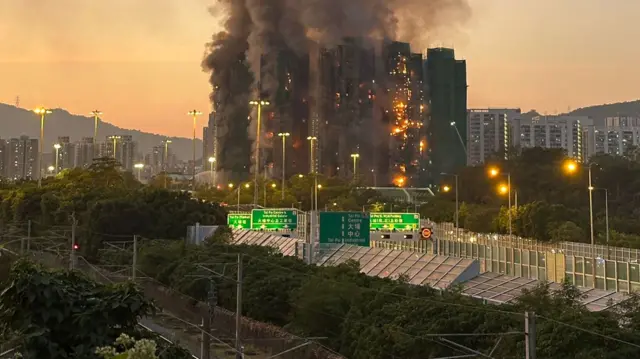 The burning Hong Kong buildings are shown behind a highway and train line as dusk descends.