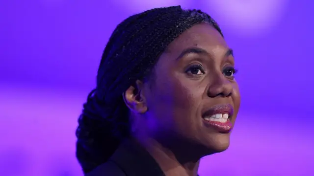 Kemi Badenohc, a black woman with dark hair in a braid looking away from the camera with a blue and purple background.