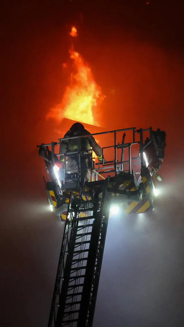 Firefighter on a cherry-picker with flames visible behind