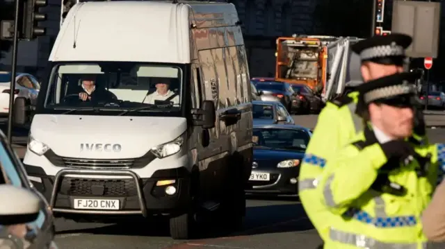A prison van ariving at Liverpool Crown Court, with cars in the background and two police officers in hi-vis in the foreground.