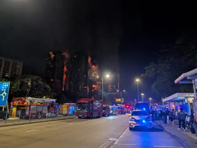 Buildings on fire in the background, several fire engines parked on the road and the side of the road