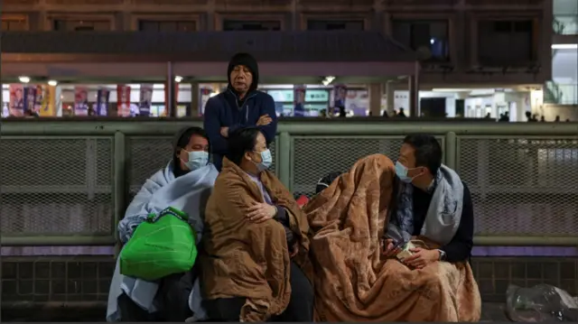 A woman and three men sit down on the side of a road outside the burning buildings. The group are all wearing face masks and are using brown and blue duvets to protect themselves from the cold