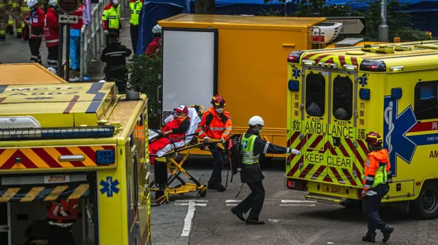 Rescue workers transport a victim on a stretcher near a fire at Wang Fuk Court. There are two ambulances and lots of emergency workers in shot.