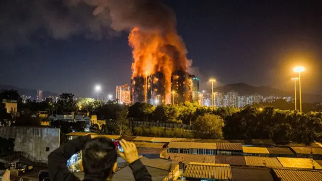 An onlooker takes photos as a major fire engulfs several apartment blocks at the Wang Fuk Court residential estate in Hong Kong's Tai Po district on 26 November 2025.