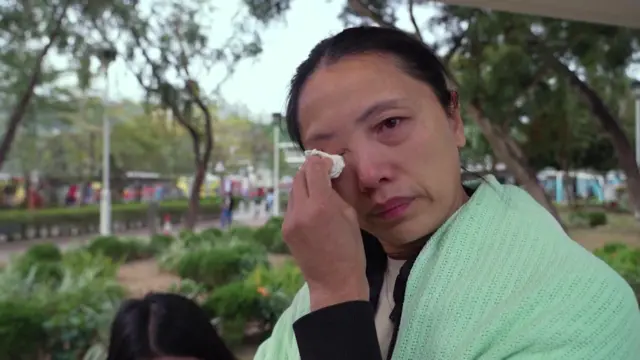 Mrs Wan stood outside. She's covered in a blanked and is wiping tears out of her eye with a tissue, looking at the camera.
