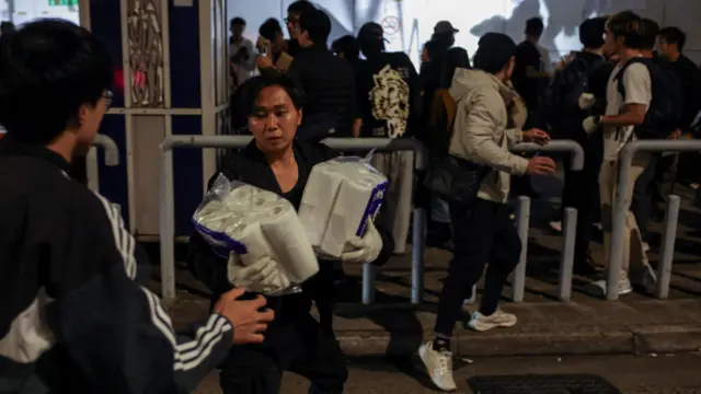People pass toilet rolls between them as crowds of people await in the background