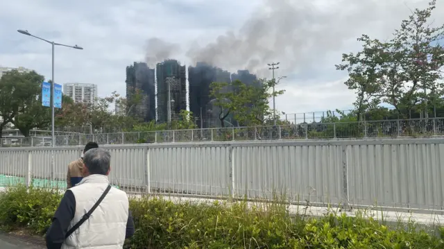 An elderly man in a white puffer vest looking into a distance, where smoke is rising from a cluster of apartment blocks