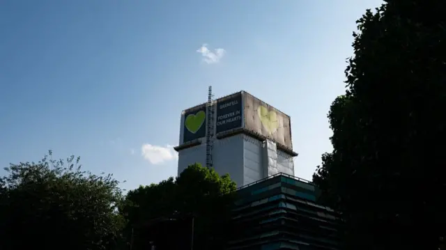 Grenfell Tower seen through trees. The cover is covered in scaffolding and a banner which says 'Grenfell. Forever in our hearts'