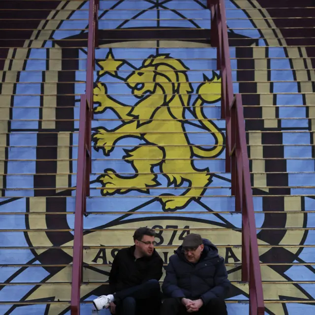 Villa fans sit in front of a mural near the ground