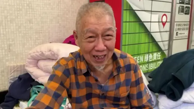 An older man sits on the floor of a shopping centre, surrounded by blankets and what is clearly a makeshift sleeping situation. He's wearing a checked shirt.