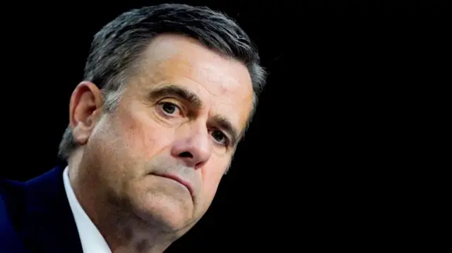 Ratcliffe in suit and tie, with serious expression, against a black background