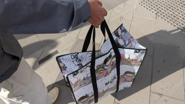 A big carrier bag held by Mrs Cheung, who wears a grey top and white trousers