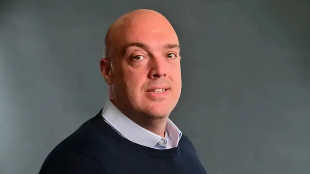 Ian Walker looks at the camera in a studio wearing a navy sweater